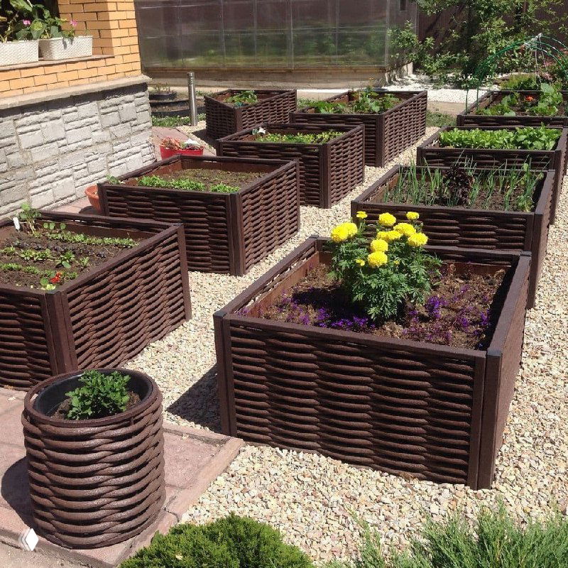 Набор квадратных грядок и кашпо из полимерной лозы на гравийной площадке; видна аккуратная расстановка контейнеров и клумб для цветов и овощей.