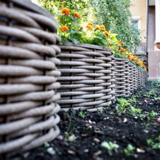 Ближний план низкого плетёного бордюра из полимерной лозы вокруг цветочной клумбы; отчетливо видна текстура материала и способ соединения витков.