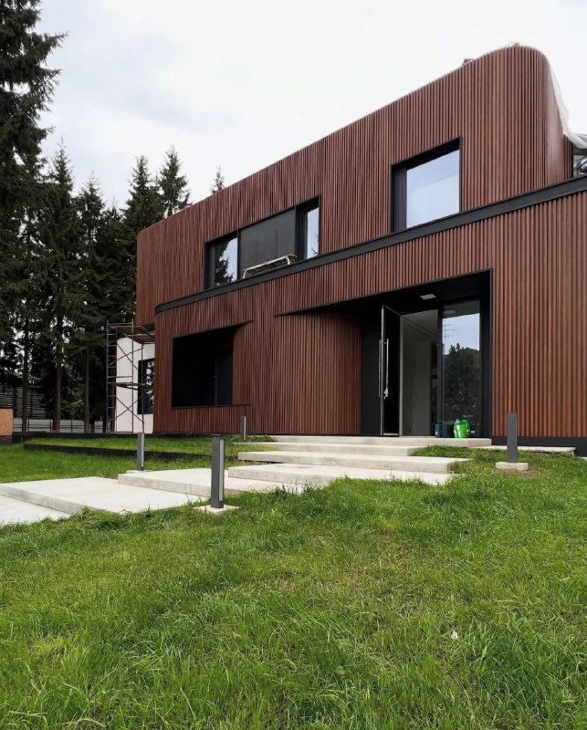 Фасад дома из брусковых ДПК, крупный план двухэтажной части с большими окнами и сочной зеленью вокруг, акцент на вертикальные рейки.