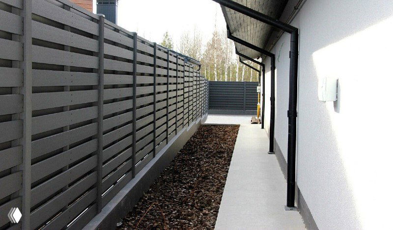 Забор из ДПК серого цвета вдоль узкого прохода у дома; показывает ровную поверхность, отсутствие необходимости в покраске и аккуратную сборку.