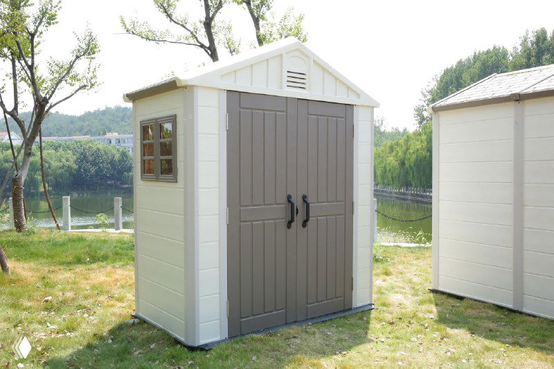 Крупный план пластикового хозблока с коричневыми дверями и боковым окном, установлен на травяном участке рядом с набережной.