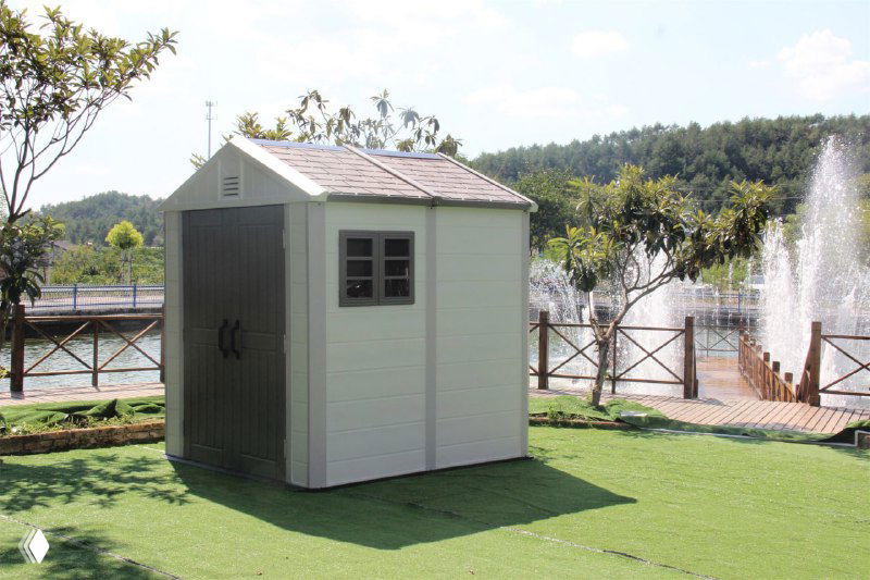Одиночный светло-серый пластиковый сарай на декоративной лужайке у фонтана — вид спереди и сбоку, демонстрация габаритов модели.