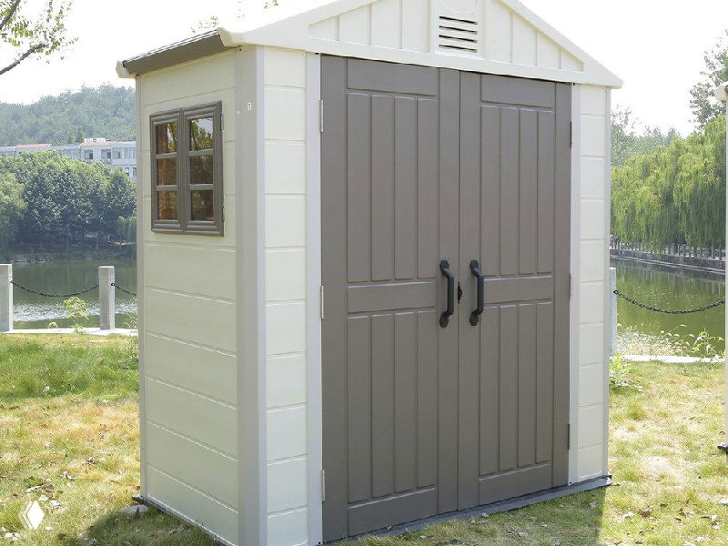 Компактный пластикoвый хозблок с дверью и окном на газоне возле водоёма, практичное решение для инструментов и хранения вещей.
