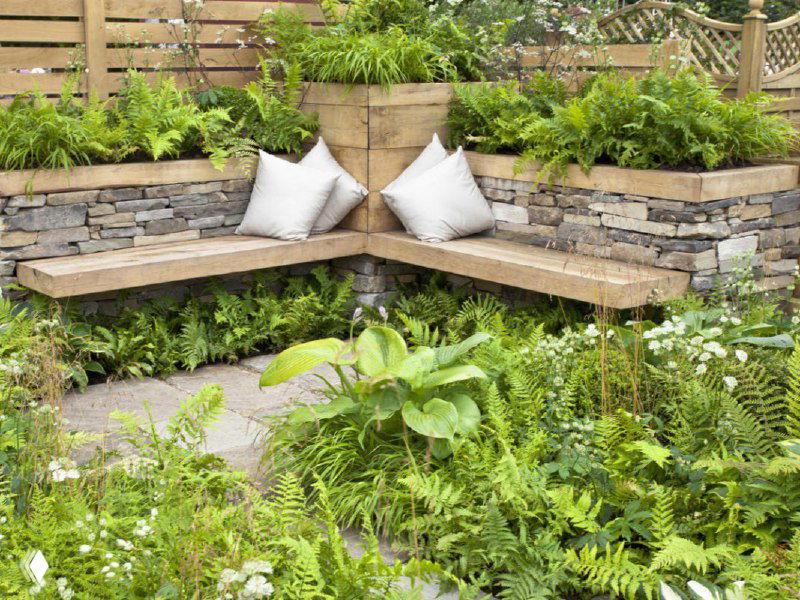 Угловая скамейка у каменной подпорной стенки в окружении декоративных растений и подушек — уютный уголок для отдыха на участке.