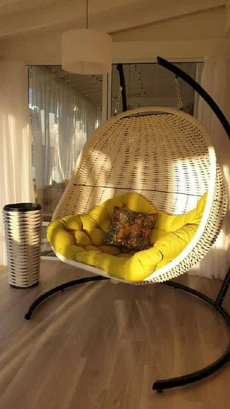 Подвесное плетёное кресло-«яйцо» из полимерной лозы внутри комнаты, яркая жёлтая подушка и тёплое солнце, лампа рядом.