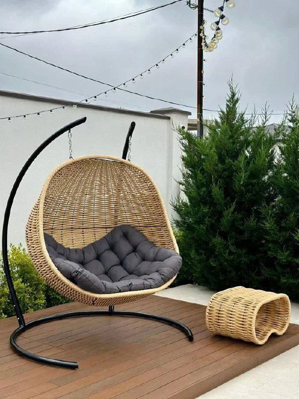 Подвесное кресло на металлической стойке из полимерной лозы на террасе, тёмно-серая подушка и маленький плетёный табурет рядом.