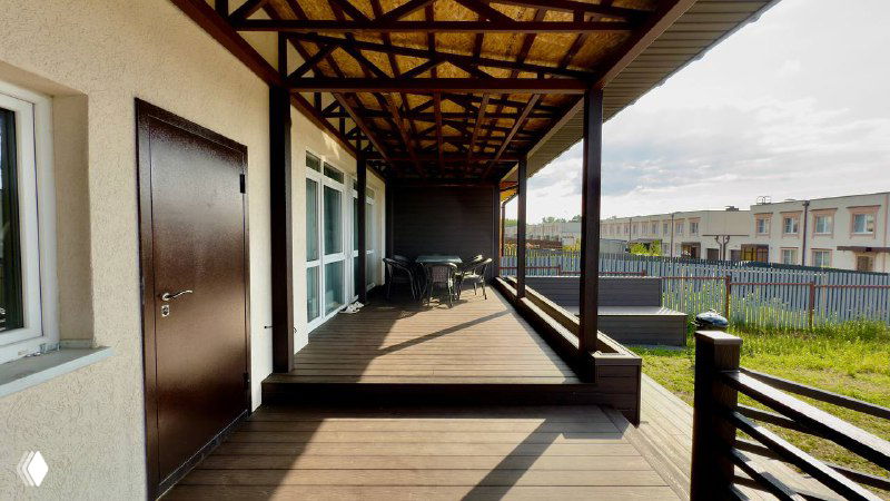 Длинный вид по настилу террасы под навесом: стол и стулья на заднем плане, навесная конструкция и однотонный композитный пол, перспектива от дома.