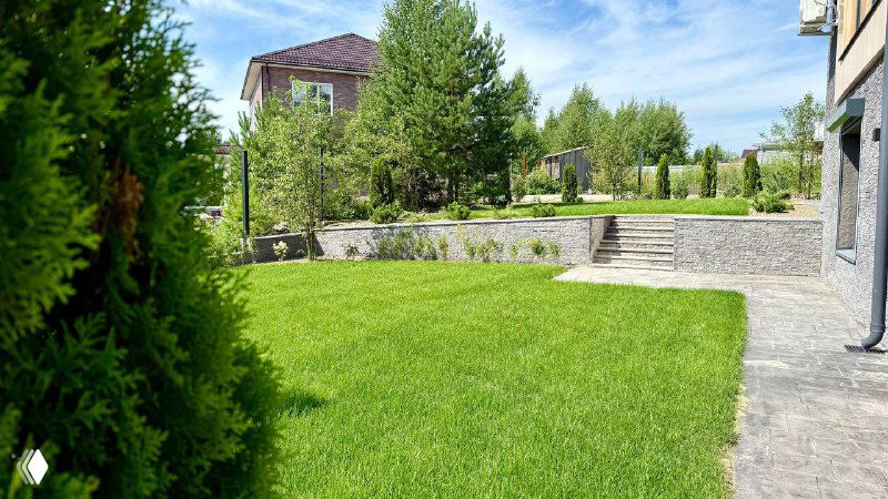 Широкий кадр ухоженного газона и приподнятой террасы, видна зелёная лужайка, деревья и каменная подпорная стена на участке.