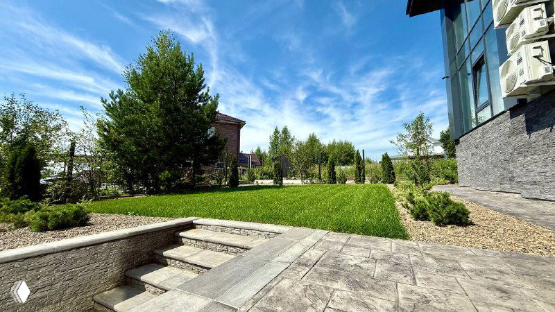 Панорамный вид на ровный рулонный газон, окружённый декоративными посадками и дорожками, чистая зелёная поверхность под синим небом.