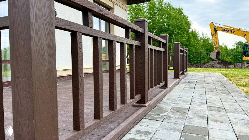 Фрагмент деревянного ограждения и поручня террасы из коричневого ДПК Polivan SINGARAJA, вид на мощёную дорожку и зелёную зону участка за ним.