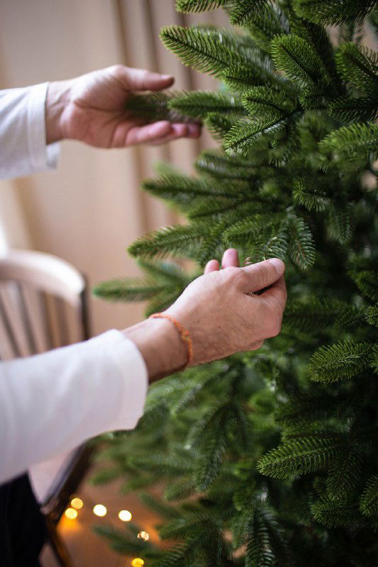 Крупный план рук, поправляющих литые веточки зелёной искусственной ёлки — видно фактуру иголок и плотность ветвления для надежной фиксации украшений.