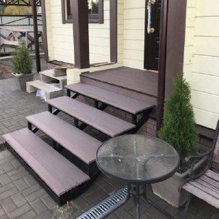 Приставное крыльцо на металлическом каркасе со ступенями из ДПК рядом с небольшой летней зоной; виден стеклянный столик и площадка у входа.