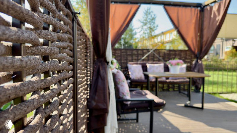 Крупный план зоны отдыха в беседке: кресла с подушками, стол и вид на плетёный забор из полимерной лозы — уют и приватность участка.