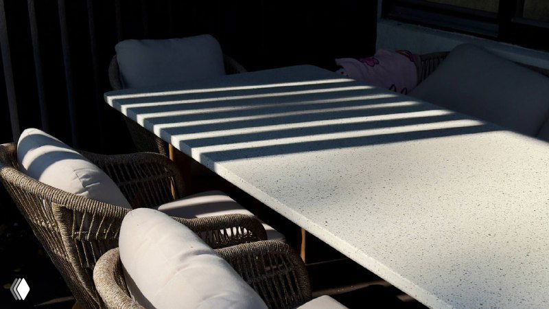 Вид на стол и диван коллекции TALARA под навесом: расстановка мебели, мягкие подушки и игра света на поверхности столешницы