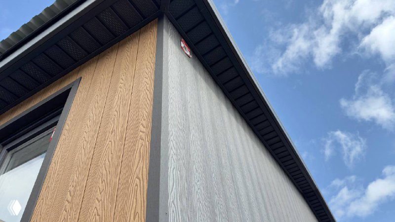 Угол модульного домика крупным планом: пересечение двух цветов фасада — современный серый и тёплый песочный, карниз и стыки панелей под небом.