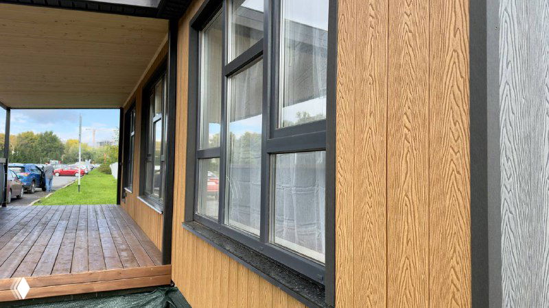 Ракурс фасада вдоль террасы: окна в тёмной раме, сочетание двух оттенков доски Polivan SINGARAJA и демонстрационная площадка выставки на заднем плане.