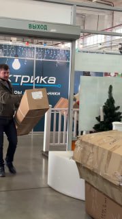 Сотрудник магазина несёт коробку с ёлкой по торговому залу LetoMarket; на фото видны стеллажи, упаковки и общий складской уголок.