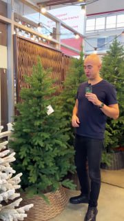 Вертикальное фото: продавец стоит рядом с большой пушистой ёлкой в плетёной корзине — крупный план ассортимента магазина LetoMarket.