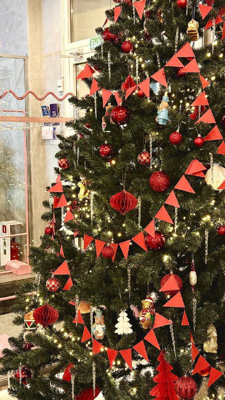 Фрагмент ёлки с бумажными гирляндами и красными шарами; видны ветви, мерцающие огоньки и часть оформления окна кафе, создающее праздничное настроение.