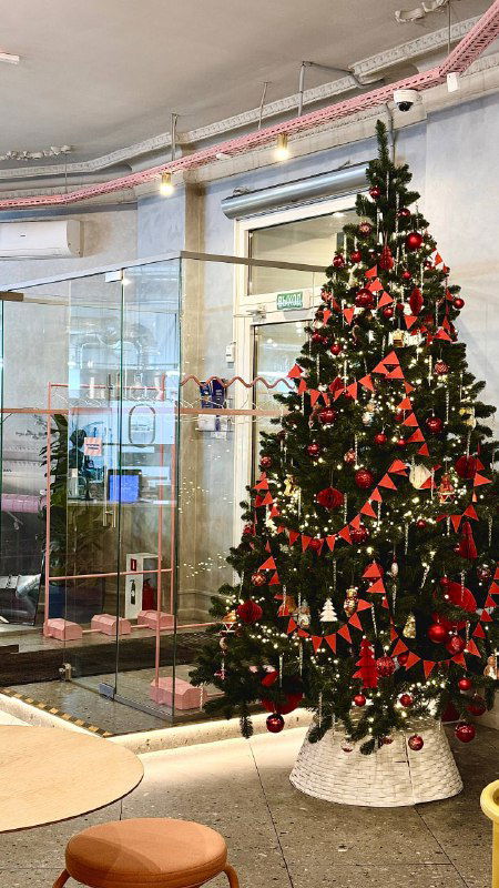 Высокая украшенная ёлка рядом со стеклянной перегородкой кафе: огни, игрушки и мягкое освещение формируют тёплый праздничный уголок для посетителей.