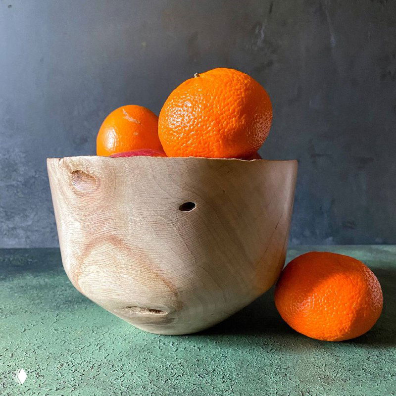 Резная деревянная чаша из светлой древесины (возможно граб) на зелёно-сером столе, рядом апельсины — натюрморт.