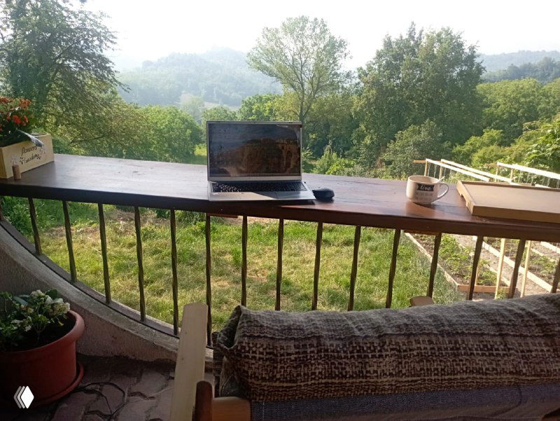 Рабочее место на балконе с ноутбуком и видом на зелёную долину — типичный образ жизни диджитал-номада в Италии, видна столешница и кресло.