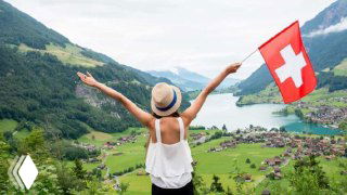 Путешественница в шляпе с швейцарским флагом на фоне озера, горной долины и деревни — символ жизни, работы и переезда в Швейцарию.