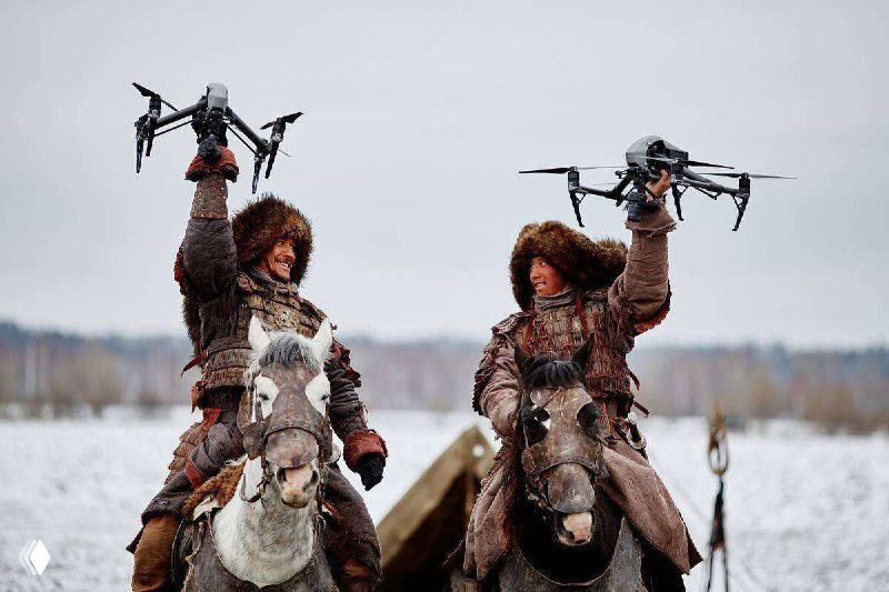 Два всадника в зимней одежде на лошадях держат над головами летательные дроны на заснеженном поле — сцена на фоне серого неба.
