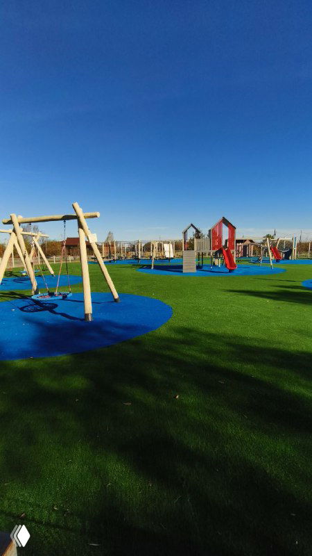 Яркая детская площадка с качелями и игровыми конструкциями на мягком покрытии в зелёной зоне базы отдыха под чистым небом.