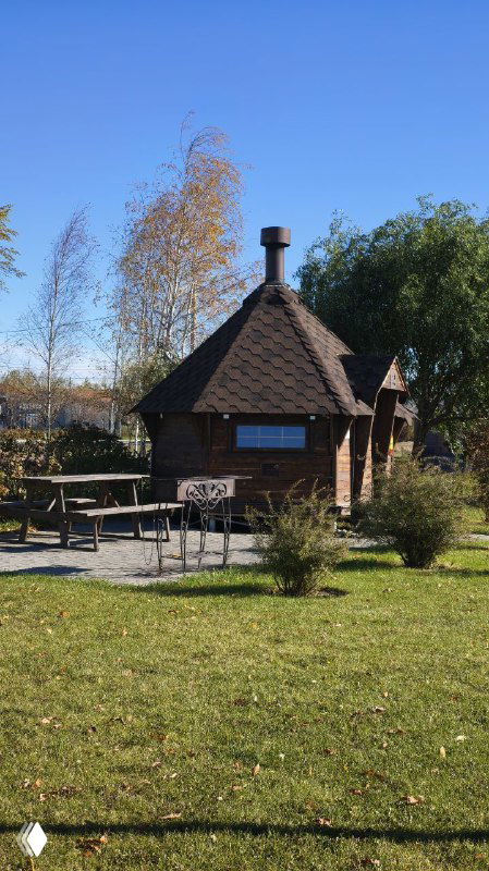 Небольшая деревянная баня с зоной для барбекю и столиками на газоне, зелёная территория вокруг и осенние деревья.