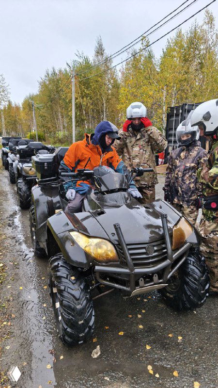 Участники и инструкторы в яркой экипировке у ряда квадроциклов на асфальтовой дороге осенним днём; подготовка к групповому заезду.
