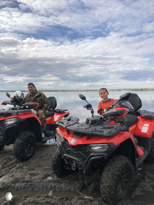 Два человека на красных квадроциклах на прибрежной грязной дороге, на переднем плане шлем и брызги грязи, солнечное небо над озером.