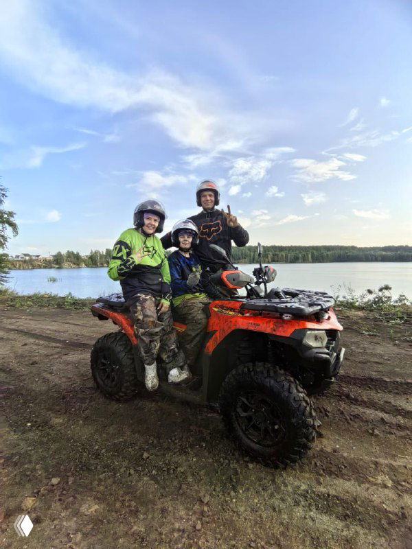 Два ребёнка в шлемах и взрослый на квадроцикле на фоне озера, улыбки и дружелюбная атмосфера семейного отдыха на свежем воздухе.