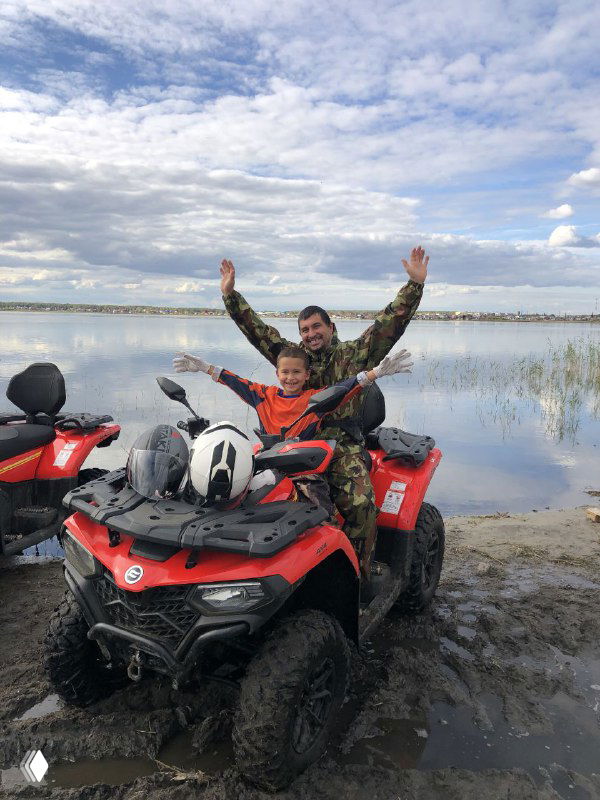 Взрослый и ребёнок радуются на красном квадроцикле у воды, руки подняты вверх, видна дорожная грязь и река на заднем плане, радостный момент.