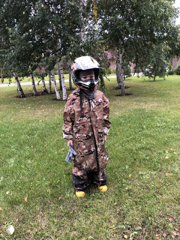 Маленький ребёнок в камуфляжном костюме и шлеме стоит на траве возле берёз, подготовка к выезду и детская экипировка в естественной обстановке.