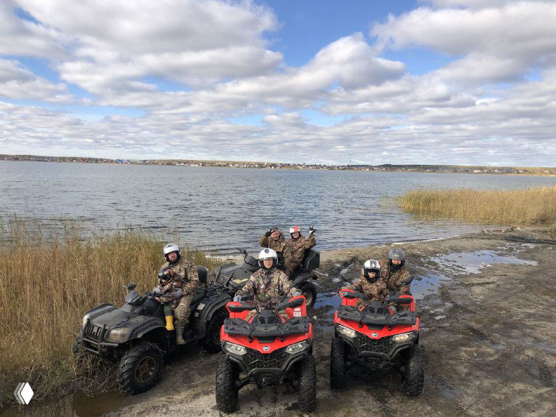 Три квадроцикла выстроены на берегу озера, рядом экипированные участники, видна водная гладь и прибрежная растительность — общий снимок группы.