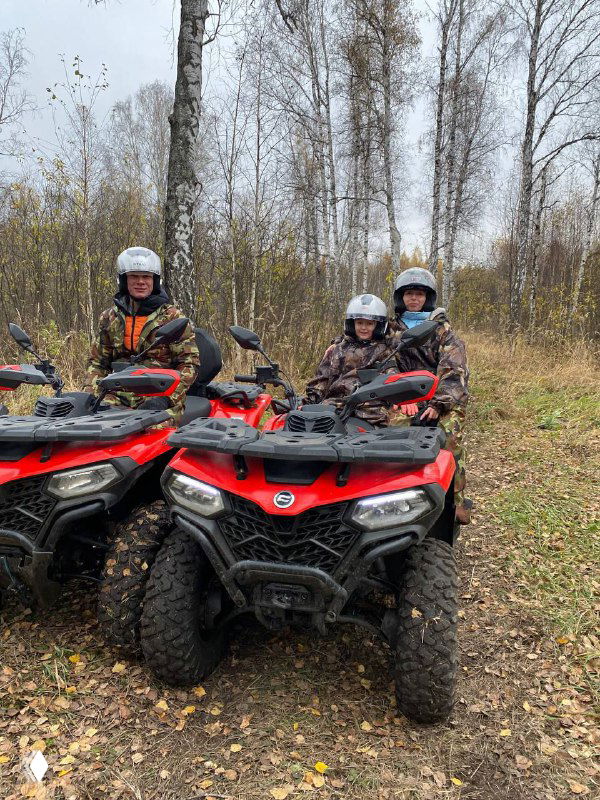 Три ребёнка в шлемах сидят на квадроциклах в лесу, видны лиственные деревья и дождливо-осенняя погода, безопасная организация детского катания.