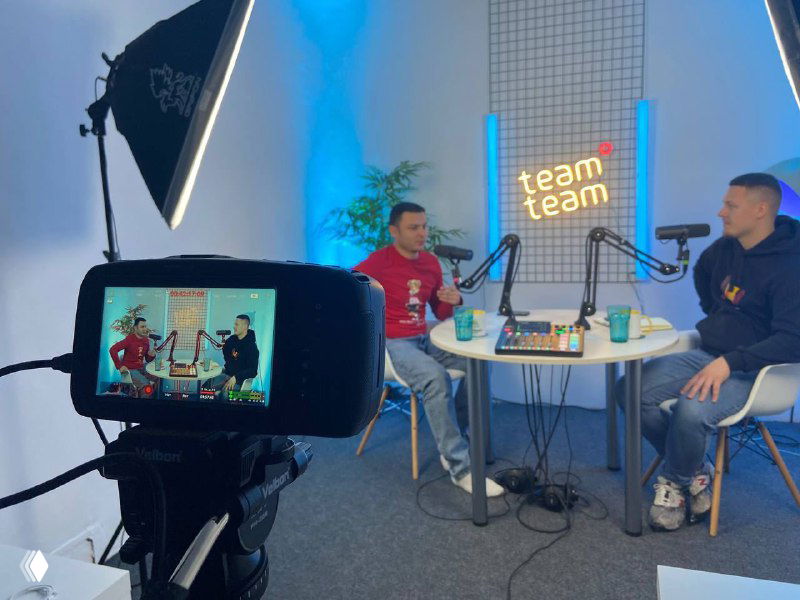 Запись подкаста в студии: два ведущих сидят за столом с микрофонами, в переднем плане камера на штативе, видна неоновая вывеска и студийное освещение.