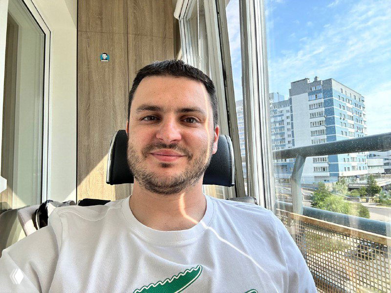 Портрет автора на балконе во время видеозвонка: мужчина сидит у окна, за ним балконная рама, вид на жилые дома и небо.