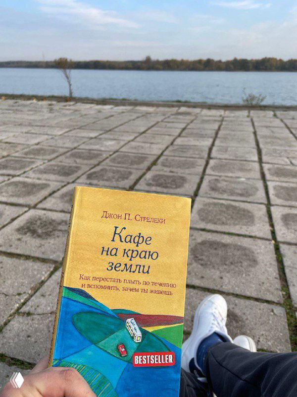 Книга «Кафе на краю земли» на плиточной набережной у воды; в кадре видны ноги в кроссовках, создавая атмосферу прогулки и раздумий автора.