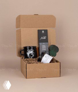 Открытая крафтовая коробка с корпоративным подарочным набором ручной работы: кружка, плитка шоколада, баночка и декоративный элемент на нейтральном фоне.