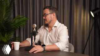 Человек в светлой рубашке разговаривает в микрофон за столом студийной записи: видна чашка, растение, софтбокс и плотные шторы на фоне.