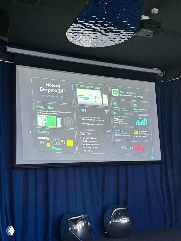 Фото с презентации: проекционный экран со слайдом новой версии «Битрикс Гравитация», занавес и два кресла на сцене, кадр в зале.