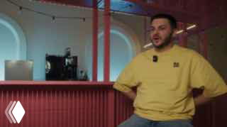 Кадр из интервью: сотрудник в жёлтой толстовке сидит в студии перед красной стойкой — фрагмент видео о биздеве и продажах, иллюстрация выпуска