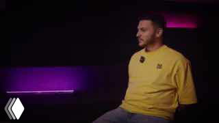 Кадр интервью: мужчина в жёлтой футболке сидит в студии с тёмным фоном и пурпурной подсветкой, разговорная сцена.