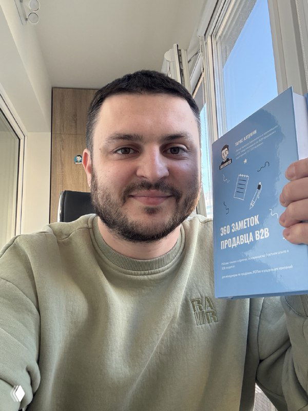 Автор улыбается и держит печатную книгу «360 заметок продавца В2В» с голубой обложкой; снимок сделан в домашнем интерьере у окна.