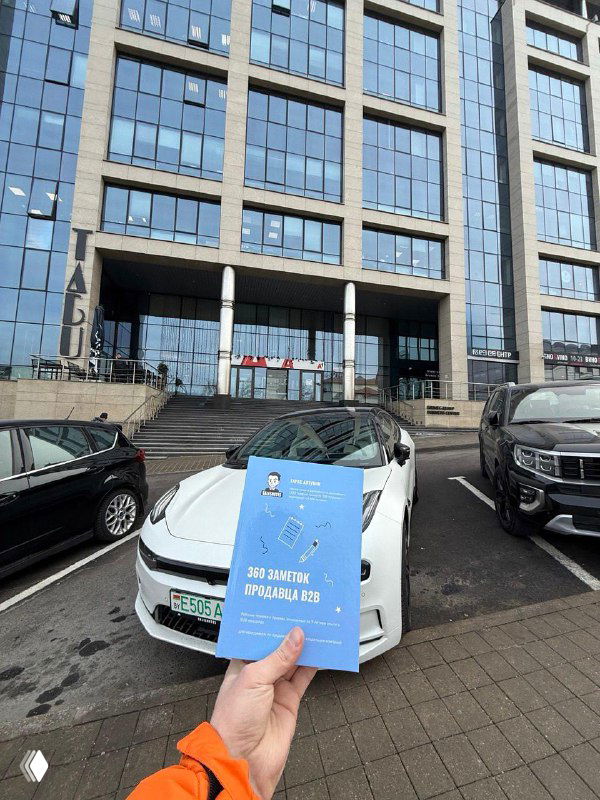 Рука держит книгу с голубой обложкой на фоне припаркованных автомобилей и стеклянного офисного здания — место, где автор писал часть заметок.