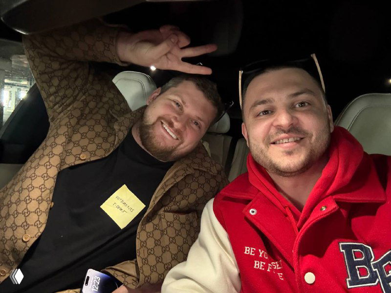 Два улыбающихся мужчины в салоне автомобиля делают селфи; один в красной толстовке, другой в коричневой куртке.