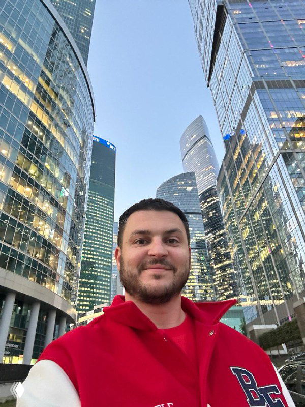 Селфи Тараса Алтунина на фоне стеклянных небоскрёбов Москва‑Сити; деловой квартал вечером, автор в красной куртке улыбается.