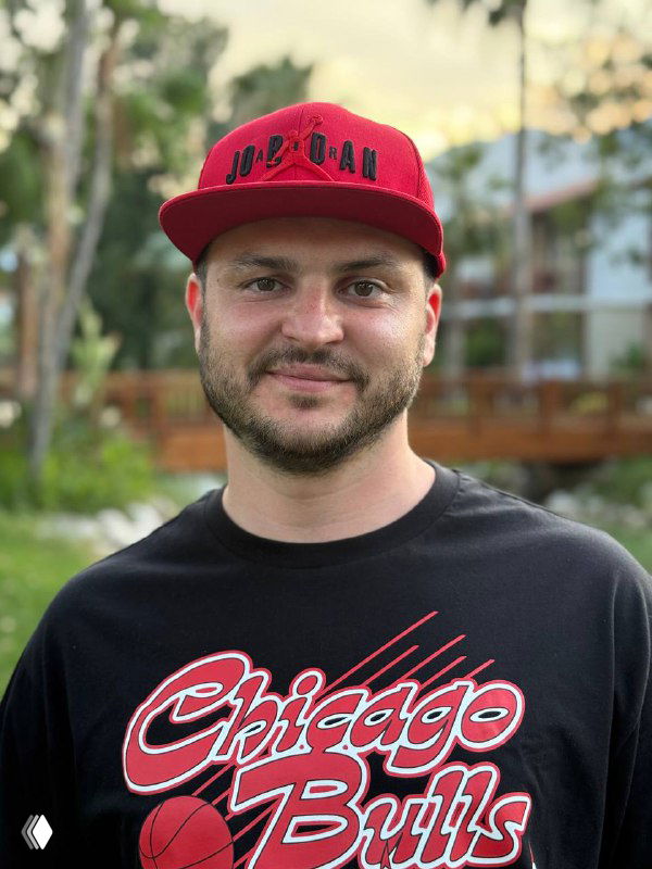 Фото автора в красной кепке и черной футболке с логотипом Chicago Bulls на фоне зелёного двора; нейтральная улыбка, прямой взгляд в камеру, повседневный образ.